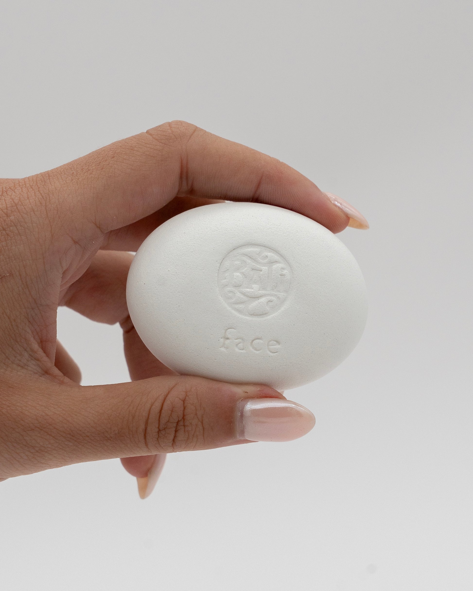 Hand holding a smooth white face stone with a subtle embossed design and the word Face Pebble, ideal for gentle exfoliation and myofascial massage.