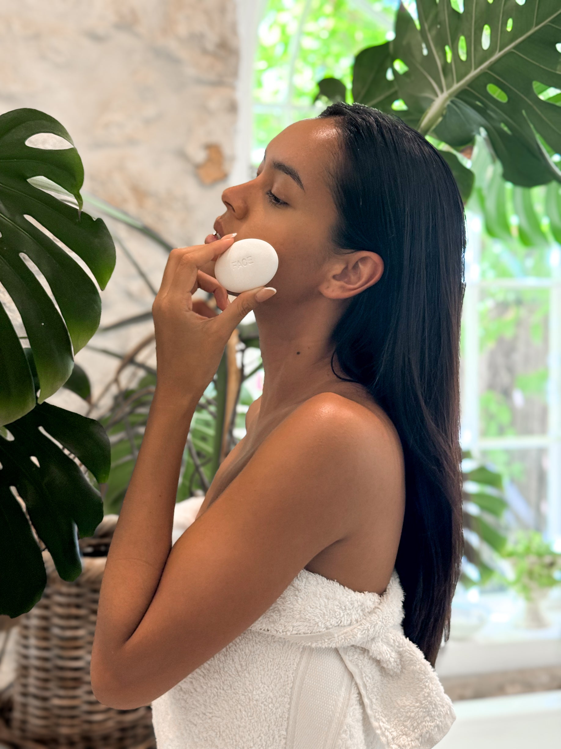 Woman with long dark hair wrapped in white towel using Face Pebble for myofascial massage and exfoliation in a bright, plant-filled room