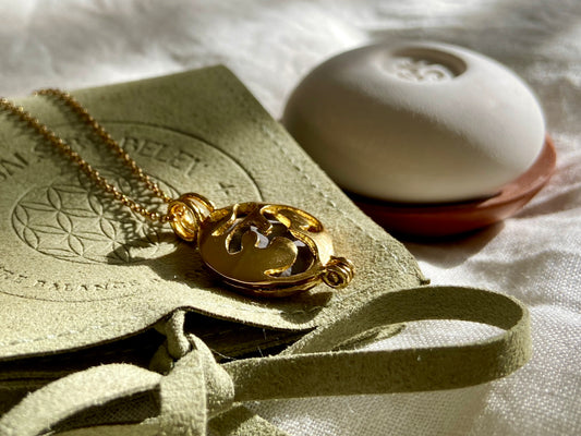 A gold-toned locket pendant necklace with a an OM design lies on a textured olive-green velvet pouch with a ribbon, next to a white ceramic pebble and wood dish on a white fabric background.