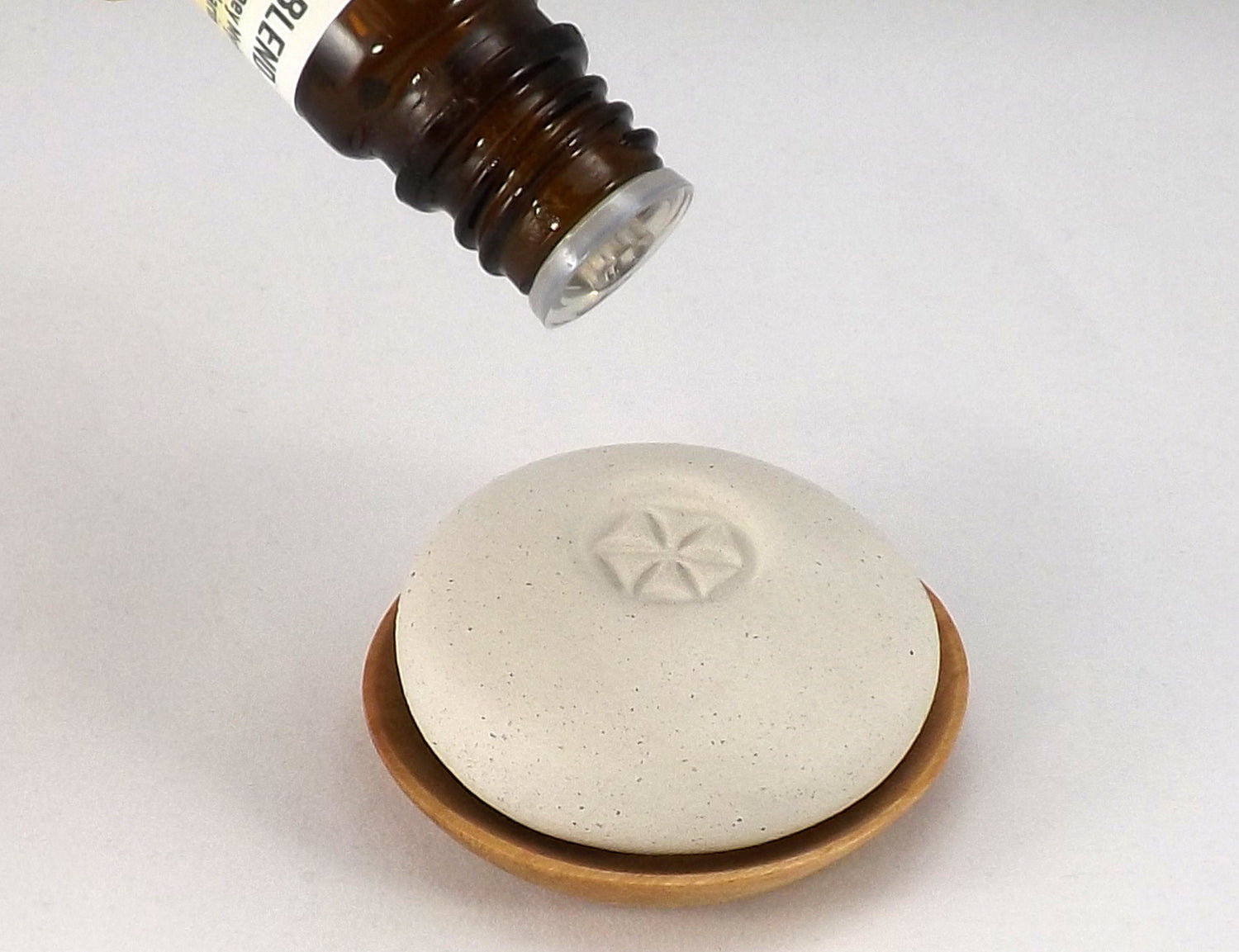 A white round pumice stone with a star-shaped engraving, resting on a small wooden dish, with a brown dropper bottle positioned above it.