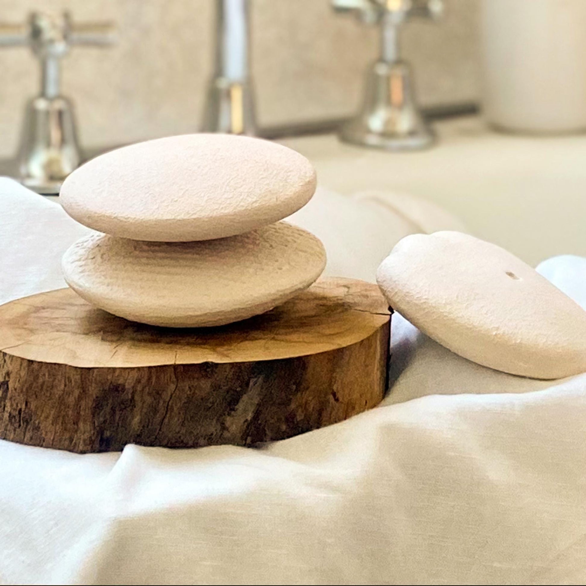 Three smooth, light-colored Body Pebble stones arranged on a wooden slab and white fabric, with blurred bathroom fixtures in the background, suggesting a calming spa setting.